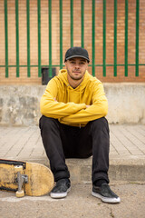 Young male in a yellow hoodie and cap with a skateboard sitting on the ground