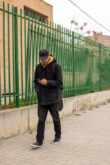 Young male with a black coat and cap walking on the street looking at the phone
