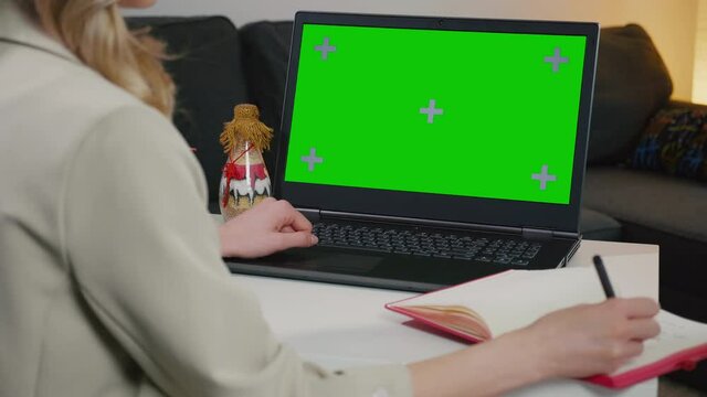 Business woman at home working on a laptop with green screen chroma key display. Working with books and computer, searching scientific article or educational information, attending online course.