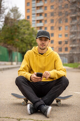 Young caucasian male from Italy in a yellow hoodie and black cap sitting on a skateboard with a smartphone