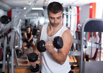 Muscular young man doing exercises with dumbbells at gym