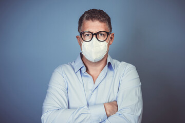 portrait of man with gray hair and blue shirt with mouth nose mask and black glasses during Corona pandemic in front of blue background