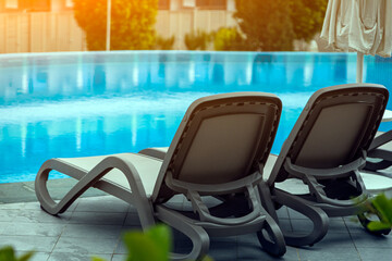 Obraz na płótnie Canvas A pair of comfortable gray sun loungers set against the backdrop of a beautiful transparent pool and hotel. The concept of a relaxing seaside vacation for two