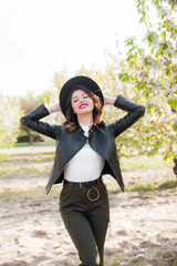 Fototapeta premium Lovely young girl in a black hat and a fashionable black leather jacket in spring in a blooming cherry garden in sunny weather with a blooming cherry branch in her hands. Spring and Fashion 