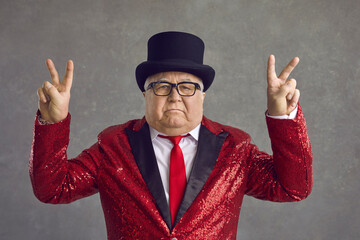 Portrait of mature millionaire on gray background. Funny rich old man in spectacles, bling fancy red sequin jacket and black top hat doing peace sign and looking at camera with serious face expression
