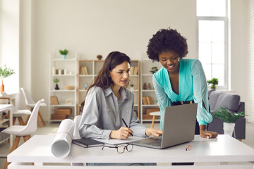 Two architects or interior designers working in the office together. Young women using laptop computer, doing Internet research, creating digital drafts, choosing new designs and making project plan