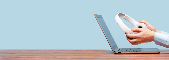 Side view of slim laptop and hands with wireless headphones on grey desk. Blue background. Distant learning. working from home, online courses or support. Audio podcast. vlogger or blogger banner