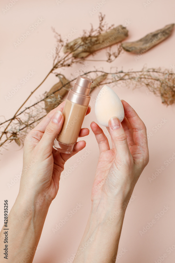 Wall mural Female Hands holding makeup liquid found or concealer and make up beuty blender or egg sponge on trendy neautral beige earth tones background, Beauty salon flyer, banner, make up artist concept