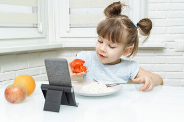 kid girl eating food and watching cartoons on a smartphone. Organic food and vegetables on a plate. A way to feed children