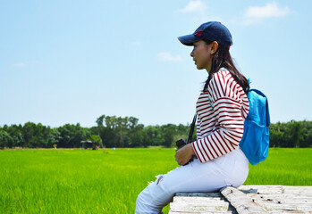 A woman with a blue backpack sits on a wooden bridge on a clear day.