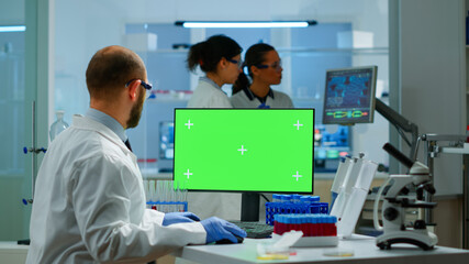 Man researcher looking at chroma key display in modern equipped lab typing on computer. Team of microbiologists doing vaccine research writing on device with green screen, isolated, mockup display.