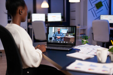 African business woman discussing with partners about deadline for marketing project during online videocall conference. Entrepreneur working on laptop computer in corporate office late at night