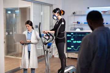 Team of professional doctors measuring health conditions of woman athlete in science sport laboratory. Physician using laptop controling EKG data, muscle endurance, heart rate psychological resistance