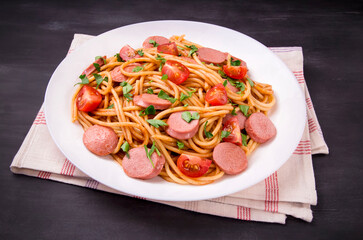 Pasta spaghetti with cherry tomatoes, fried sausage and parsley  in white plate on a black  background.