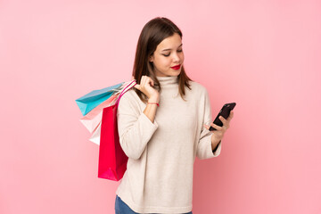 Teenager girl isolated on pink background holding shopping bags and writing a message with her cell phone to a friend