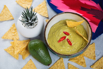 Bowl of guacamole dip sauce and corn chips, elevated view on a light-blue stone surface with cactus and mexican poncho