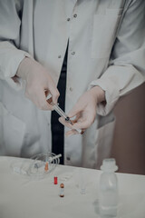 Female hands in white medical gloves drawing medicine from ampoule into a syringe, next to upright ampoule with medicine on white acrylic table.