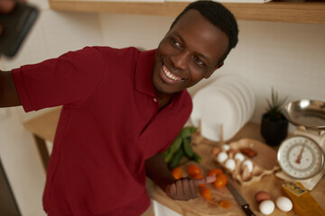 Happy attractive young black male standing in kitchen with eggs and vegetables on counter pointing, making thumbs up gesture, cooking breakfast taking selfie using mobile phone, smiling broadly