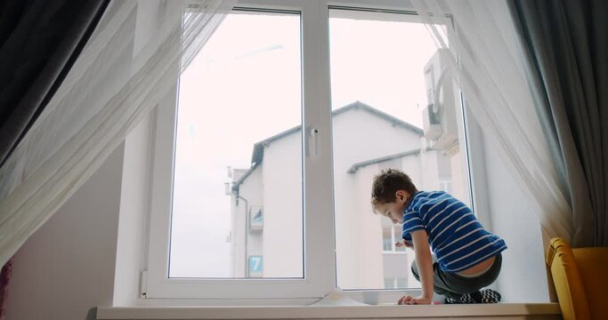 Little boy with curly hair draws on the window at home with crayons. 