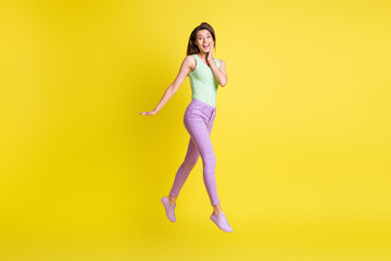 Fototapeta na wymiar Photo of lady jump go hand face wear green top purple pants sneakers isolated yellow color background