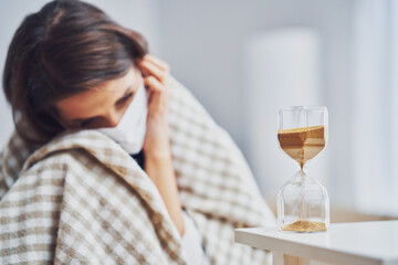 Adult woman in mask feeling sick at home with hourglass Covid-19 concept