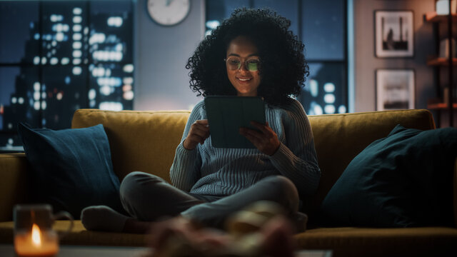 Beautiful Authentic Latina Female Sitting On Couch Sofa In A Cozy Dark Living Room In The Evening And Using Tablet Computer At Home. She's Browsing The Internet And Checking Videos On Social Networks.