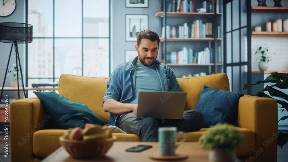 Aufkleber Handsome Caucasian Man Working on Laptop Computer while Sitting on a Sofa Couch in Stylish Cozy Living Room. Freelancer Working From Home. Browsing Internet, Using Social Networks, Having Fun.