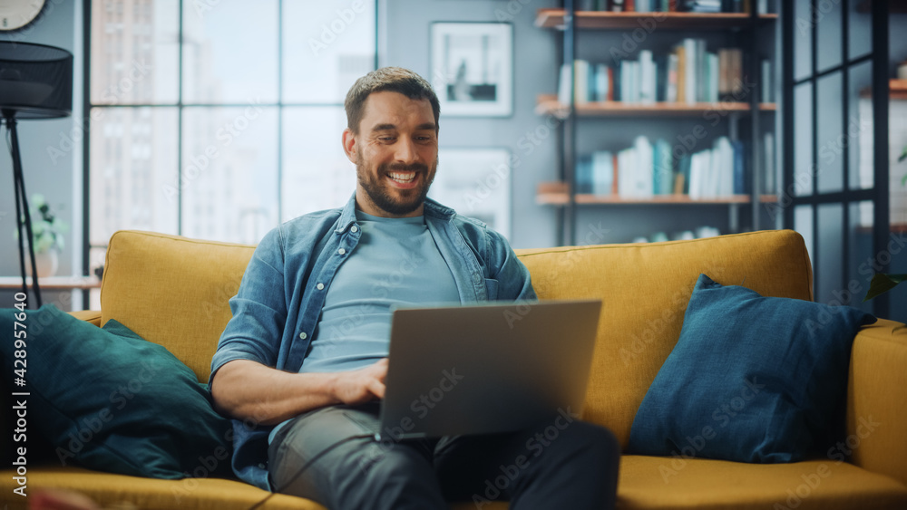 Aufkleber Handsome Caucasian Man Working on Laptop Computer while Sitting on a Sofa Couch in Stylish Cozy Living Room. Freelancer Working From Home. Browsing Internet, Using Social Networks, Having Fun.