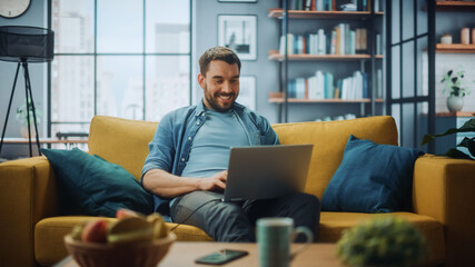 Handsome Caucasian Man Working on Laptop Computer while Sitting on a Sofa Couch in Stylish Cozy...