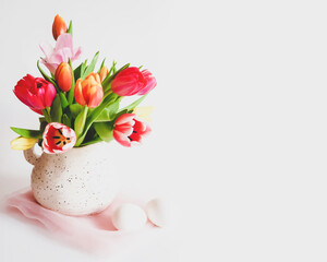Easter composition with multicolored tulips in a jug, white eggs on a white background with copy space. Preparing for Easter. Spring holiday mood. Flower still life, horizontal orientation