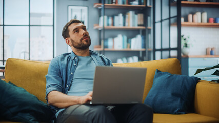 Handsome Caucasian Man Working on Laptop Computer while Sitting on a Sofa Couch in Stylish Cozy...