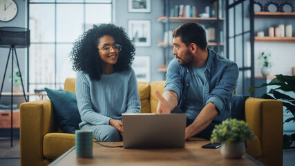 Diverse Multiethnic Couple are Sitting on a Couch Sofa in Stylish Living Room and Choosing Items to Buy Online with Laptop Computer. Friends or Colleagues are Discuss Work Projects.