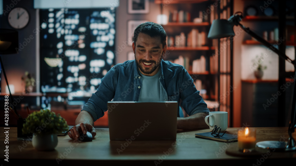 Poster Handsome Caucasian Man Working on Laptop Computer while Sitting at a Desk in Dark Cozy Living Room in the Evening. Freelancer Working From Home. Browsing Internet, Using Social Networks, Having Fun.