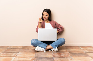 Young mixed race woman with a laptop sitting on the floor making Italian gesture