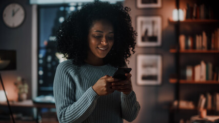 Portrait of a Beautiful Authentic Latina Female in a Stylish Dark Cozy Living Room Using Smartphone at Home. She's Browsing the Internet and Checking Videos on Social Networks and Having Fun.