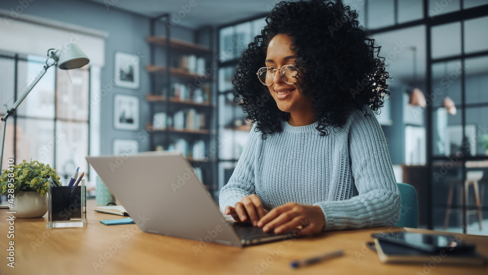 Poster beautiful authentic latina female with afro hair sitting at a desk in a cozy living room and using l