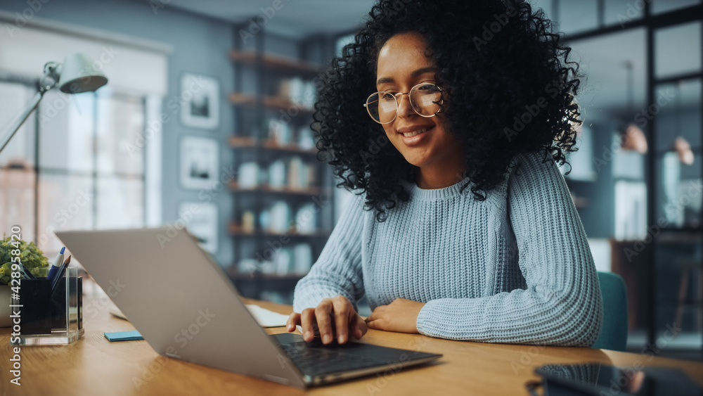 Poster beautiful authentic latina female with afro hair sitting at a desk in a cozy living room and using l