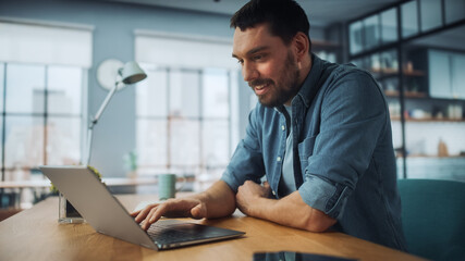 Fototapeta na wymiar Handsome Caucasian Man Working on Laptop Computer while Sitting on a Sofa Couch in Stylish Cozy Living Room. Freelancer Working From Home. Browsing Internet, Using Social Networks, Having Fun.