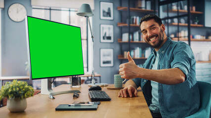 Naklejka na ściany i meble Handsome Caucasian Specialist Working on Desktop Computer with Green Screen Mock Up Display at Home Living Room. Happy Freelance Man Smiles to the Camera and Shows a Thumbs Up Hand Gesture.