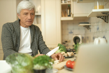 Serious concentrated retired man cooking dinner in cozy kitchen slicing fresh ingredients on cooking board, watching football game or series online on open laptop, having focused facial expression