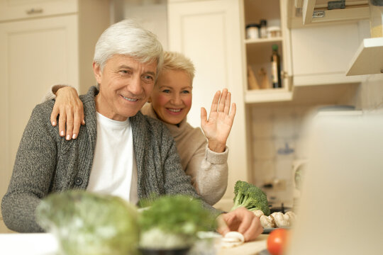 Happy Cheerful Middle Aged Couple Spending Time Together At Home Making Dinner In Stylish Kitchen Interior. Retired Woman And Man Waving Hand At Webcam While Having Video Conference Call On Laptop