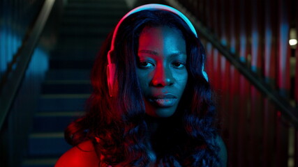 Portrait Of Black Young Girl In Headphones. She Stands On The Background Of The Stairs. Lighting In Different Colors.