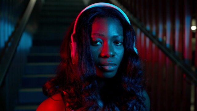 Portrait Of Black Young Girl In Headphones. She Stands On The Background Of The Stairs. Lighting In Different Colors.