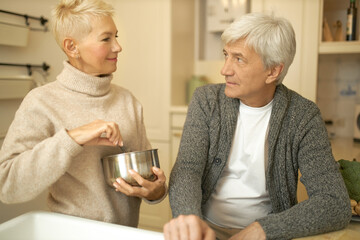People, home, cooking and leisure time. Confident mature housewife whipping eggs in saucepan using whisk having conversation with her gray haired senior husband. Retired couple making dinner