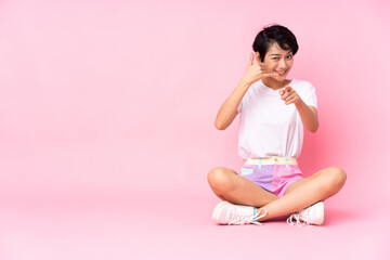 Young Vietnamese woman with short hair sitting on the floor over isolated pink background making phone gesture and pointing front