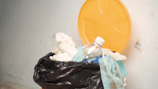Close up of healthcare worker or doctor throwing single use disposable medical face mask and gloves into filled trash can or garbage bin at hospital during coronavirus covid-19 pandemic