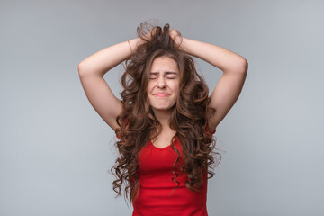 Fototapeta premium Anxious and troubled young brunette woman grab head with hands, close eyes, standing over gray background