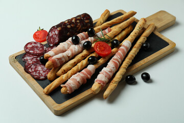 Board with grissini sticks with bacon and snacks on white background