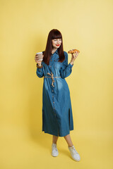 Food and drink promo ad. Full-length shot of young lady in casual trendy jeans dress and sneakers, posing on yellow background and holds delicious croissant and coffee to go