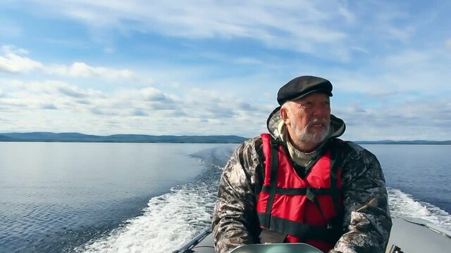 Elderly gray-haired fisherman in life jacket driving motorboat rushing along water surface of sea
